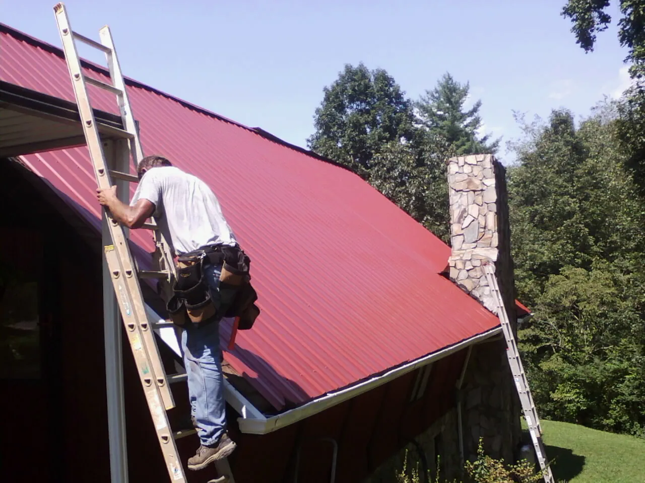 Roofer on a ladder beside a red metal roof and stone chimney — Soffit & Fascia Repair by Roofing Brothers in Tannersville